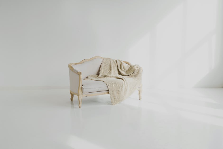 A vintage-style, cream-colored upholstered sofa with wooden decorative trim and curved legs, situated in a brightly lit, minimalist room with white walls and flooring. A beige throw blanket is casually draped over one armrest, contributing to a tidy and clean appearance. The clean, dust-free surface and natural light reflecting off the room emphasize a high standard of surface cleaning and maintenance, consistent with services offered by Belgravia Carpet Cleaning for upholstery and domestic cleaning on Elizabeth Street Belgravia.