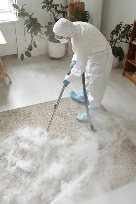 A professional cleaner dressed in a white protective suit, mask, gloves, and shoe covers is using a steam or chemical carpet cleaning machine on a light-colored, textured carpet in a bright, modern living room. The room features natural light coming through windows, with potted plants and wooden furniture visible in the background. The cleaner from Belgravia Carpet Cleaning is preparing to deep clean the carpet to enhance hygiene and restore its appearance, reflecting high-quality surface cleaning and sanitisation practices typical of domestic cleaning services aimed at maintaining a clean and fresh environment near Eaton Square SW1X, Belgravia.