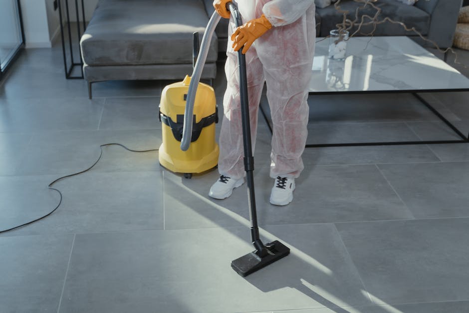 A person wearing white protective clothing and yellow gloves is performing surface cleaning in a modern living room. They are using a vacuum cleaner connected to a yellow cylinder, with a flexible hose, to deep clean large grey ceramic floor tiles that appear freshly washed and shine under natural light. The room features a grey sofa with plush cushions positioned against the wall, a glass-topped coffee table, and a decorative wooden sculpture on a side table. The room is well-lit, clean, and organized, with a neutral color palette. This cleaning is part of a professional domestic cleaning service provided by Belgravia Carpet Cleaning, focusing on maintaining hygiene and surface cleanliness.