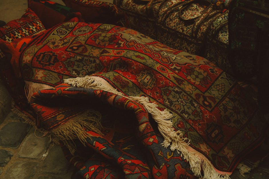 Close-up of a patterned, Persian-style rug with intricate red, orange, and beige geometric and floral designs, partially rolled or folded over, revealing its fringed edges. The rug is placed on a stone floor inside a room with dim lighting, creating a cozy, warm atmosphere. The image showcases the surface condition, highlighting the deep, detailed pattern and the clean, well-maintained appearance, consistent with professional cleaning and surface care. Belgravia Carpet Cleaning's logo or branding is not visible, but the setting aligns with their domestic cleaning services focused on deep cleaning and sanitisation of carpet and rug surfaces.