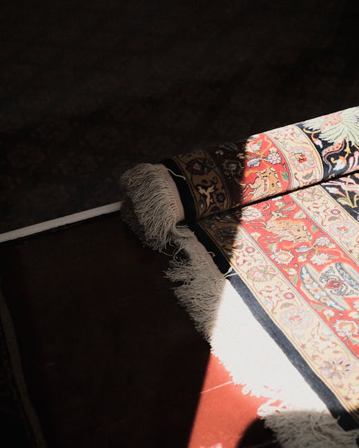 Close-up image of a traditional Persian-style rug with intricate floral patterns in shades of red, blue, and ivory, partially unrolled on a dark wooden floor in a well-lit room. The rug's edges show detailed fringes, and a white pole is visible to assist in unrolling the rug. The lighting highlights the rich colors and detailed design of the rug, indicating a clean and well-maintained surface. This image exemplifies surface cleaning and deep cleaning practices, as conducted by Belgravia Carpet Cleaning, to ensure hygiene and maintenance of domestic flooring in a residential setting.