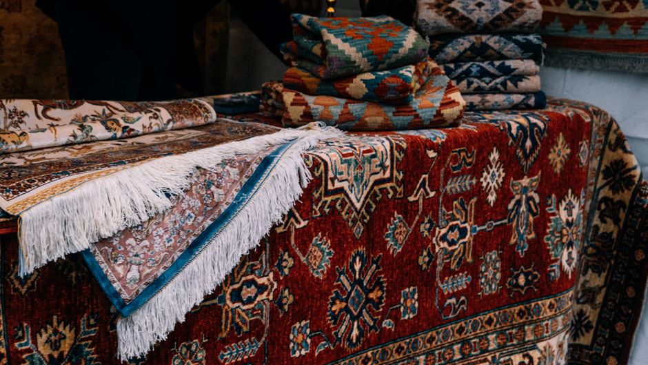 Close-up of a patterned, Persian-style rug with intricate red, orange, and beige geometric and floral designs, partially rolled or folded over, revealing its fringed edges. The rug is placed on a stone floor inside a room with dim lighting, creating a cozy, warm atmosphere. The image showcases the surface condition, highlighting the deep, detailed pattern and the clean, well-maintained appearance, consistent with professional cleaning and surface care. Belgravia Carpet Cleaning's logo or branding is not visible, but the setting aligns with their domestic cleaning services focused on deep cleaning and sanitisation of carpet and rug surfaces.