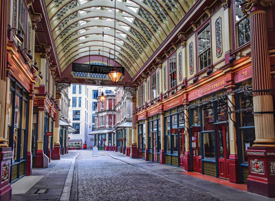 A covered shopping arcade featuring ornate architectural details in red, cream, and gold. The high, arched glass ceiling allows natural light to illuminate the cobblestone walkway below. Several storefronts with large glass windows and colorful signage line both sides, including a pizza restaurant. The storefronts are decorated with decorative columns and moldings, and the overall appearance is clean and well-maintained, reflecting professional cleaning and upkeep by Belgravia Carpet Cleaning. The scene is empty of customers, emphasizing the neatness and pristine condition of the surfaces, suited for surface cleaning and deep sanitisation services. Bright, warm lighting highlights the detailed design of the arcade's interior.
